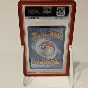 May include: A graded Pok&eacute;mon trading card in a red glittery holder, displayed on a white stand. The card's back is visible, featuring the Pok&eacute;mon logo and a Pok&eacute; Ball design. The holder has the PSA logo.