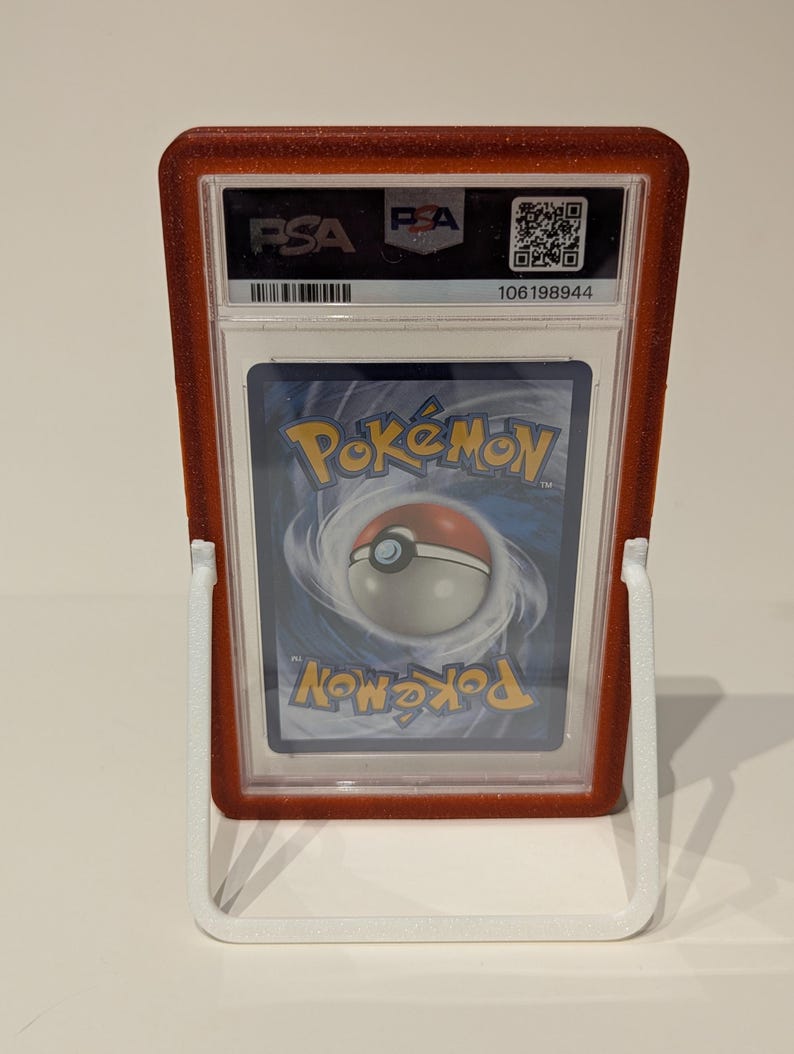 May include: A graded Pok&eacute;mon trading card in a red glittery holder, displayed on a white stand. The card's back is visible, featuring the Pok&eacute;mon logo and a Pok&eacute; Ball design. The holder has the PSA logo.