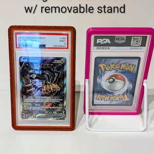 May include: Two magnetic slab cases with removable stands. One case is brown and the other is pink. The cases hold Pokemon trading cards. The text on the cases reads "Magnetic slab case w/ removable stand".