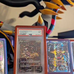 May include: A graded Pok&eacute;mon card featuring Giratina V from the Lost Origin set. The card is encased in a red glittery holder. The card's text includes "Giratina V" and "Abyss Seeking". A large, detailed Giratina figure is in the background.
