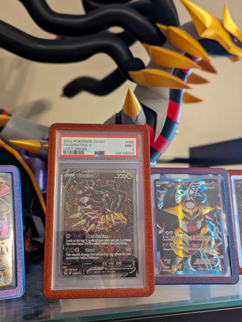 May include: A graded Pok&eacute;mon card featuring Giratina V from the Lost Origin set. The card is encased in a red glittery holder. The card's text includes "Giratina V" and "Abyss Seeking". A large, detailed Giratina figure is in the background.