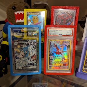May include: A collection of graded Pok&eacute;mon trading cards in protective cases. The cards are displayed in blue, red, and yellow frames. One card features a blue and white illustration of a water Pok&eacute;mon. Another card shows a red and orange Pok&eacute;mon.