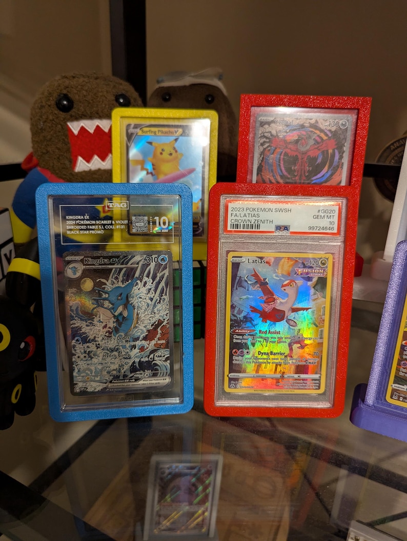 May include: A collection of graded Pok&eacute;mon trading cards in protective cases. The cards are displayed in blue, red, and yellow frames. One card features a blue and white illustration of a water Pok&eacute;mon. Another card shows a red and orange Pok&eacute;mon.