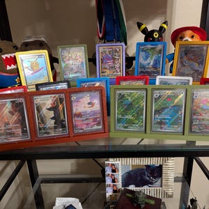 Op de afbeelding: Een display van ingelijste ruilkaarten in verschillende kleuren, waaronder rood, groen en blauw. De kaarten bevatten kleurrijke illustraties en zijn gerangschikt op een glazen plank. Pluche speelgoed en andere decoratieve items zijn ook zichtbaar.