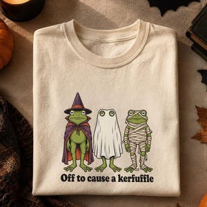 Może przedstawiać: Kremowy t-shirt z grafiką przedstawiającą trzy zielone żaby w kostiumach na Halloween: jedną jako czarownicę w fioletowym kapeluszu i pelerynie, drugą jako ducha pod białym prześcieradłem oraz trzecią jako mumię owiniętą bandażami. Napis głosi, Off to cause a kerfuffle.