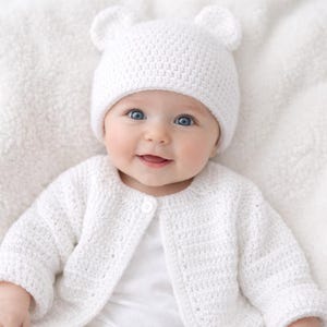 May include: A baby wearing a white crochet bear hat with two small ears and a matching cardigan. The cardigan fastens with a single button. The baby is smiling. The background is a white, textured surface.