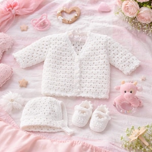 May include: A white crochet baby cardigan, bonnet, and booties set. The set includes a button-down sweater, a bonnet with a tie, and small shoes. A pacifier, a wooden heart, and a pink stuffed animal are also in the image.