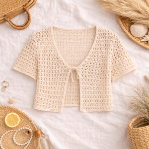 May include: A cream-coloured, open-front crochet cardigan with short sleeves and a tie closure. The lightweight knitwear features a delicate, open weave design. The cardigan is displayed on a white surface, with other accessories in the background.