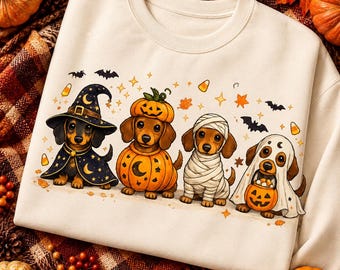 May include: A cream-colored crewneck sweatshirt featuring a graphic of four dachshunds dressed in Halloween costumes. The dogs are depicted as a wizard, a pumpkin, a mummy, and a ghost holding a jack-o'-lantern bucket, surrounded by bats and stars.