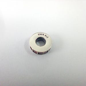 May include: A silver-colored charm with a circular design. The charm has a band of small, dark red stones around the middle. The text "S925 ALE" is stamped on the top surface. The charm is likely for a bracelet or necklace.