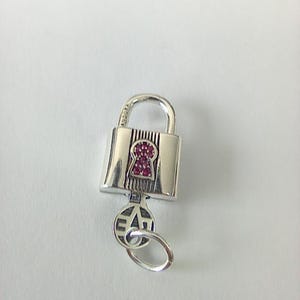 May include: Silver padlock charm with a key. The lock has a keyhole filled with small, red gemstones. The key is attached to the lock with a ring and has the word "LOVE" engraved on it. The charm is on a white background.