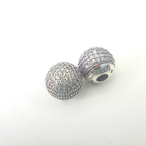 Puede incluir: Dos abalorios Pandora de plata. Cada abalorio es esférico y está cubierto de pequeñas piedras transparentes y brillantes. Un abalorio tiene el logotipo de Pandora. Los abalorios están sobre un fondo blanco.