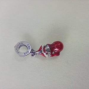 May include: A red and silver charm in the shape of a baby's dummy. The charm features a glossy red enamel finish on the dummy part and silver-coloured metal detailing. The charm is engraved with "Pandora" and "ALE S925" on the silver ring.