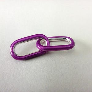 以下が含まれることがあります： 2つの連結した楕円形のチャーム。チャームは鮮やかな紫で、内側はシルバーです。チャームは、ネックレスやブレスレットのリンクなど、ジュエリーのコンポーネントである可能性があります。背景はプレーンな白です。