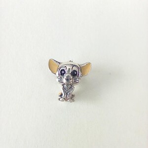Puede incluir: Un charm de color plateado que representa a un cachorro de león de dibujos animados con orejas color canela y una cara detallada. El charm está sentado con las patas delanteras. El charm es probablemente de metal y tiene un diseño detallado.