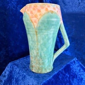 Puede incluir: Una jarra de cerámica con cuerpo cónico y superficie texturizada. La parte superior es de un suave color melocotón con un patrón moteado, mientras que la sección inferior es de un verde espuma de mar apagado. Incluye un asa angular lateral.