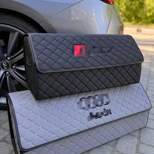 May include: Two car boot organisers. The top one is black with a diamond pattern and a red square logo. The bottom one is grey with a diamond pattern and a blurred logo. Both have a rectangular shape.