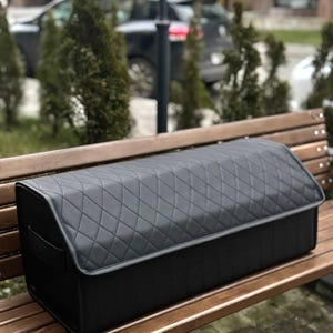 May include: A black, quilted car trunk organizer with a rectangular shape and a hinged lid. The organizer is made of a durable material and features a diamond pattern. It is designed to keep the car trunk organized and tidy.