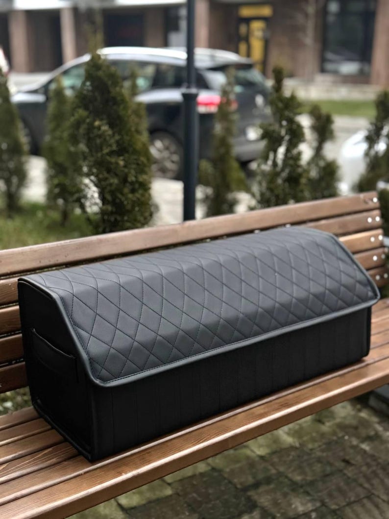 May include: A black, quilted car trunk organizer with a rectangular shape and a hinged lid. The organizer is made of a durable material and features a diamond pattern. It is designed to keep the car trunk organized and tidy.