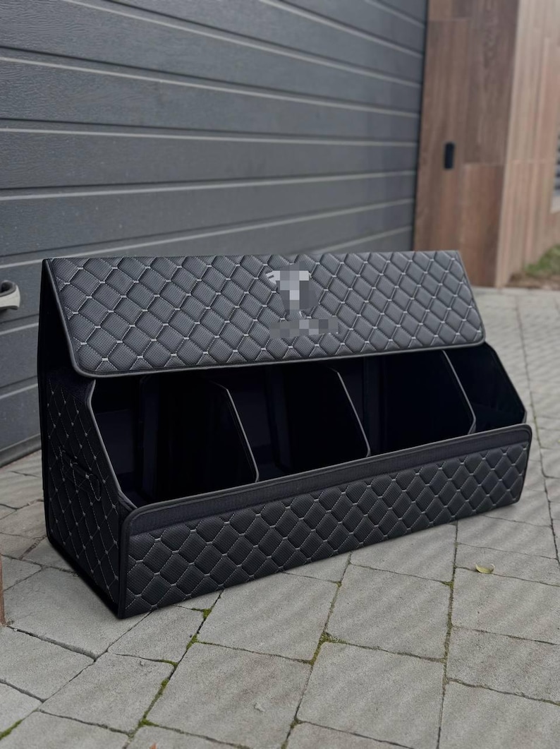 May include: Black car trunk organizer with a quilted diamond pattern and white stitching. The organizer has three compartments and a hinged lid. The exterior is a solid black color.
