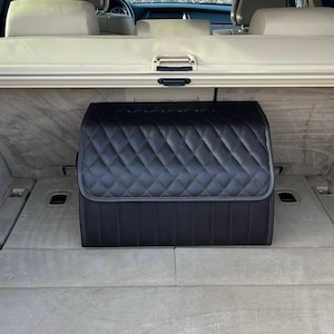 May include: Black quilted car trunk organizer. The organizer is rectangular with a diamond pattern and a top flap. It is designed to fit in the trunk of a vehicle. The interior of the car is visible in the background.