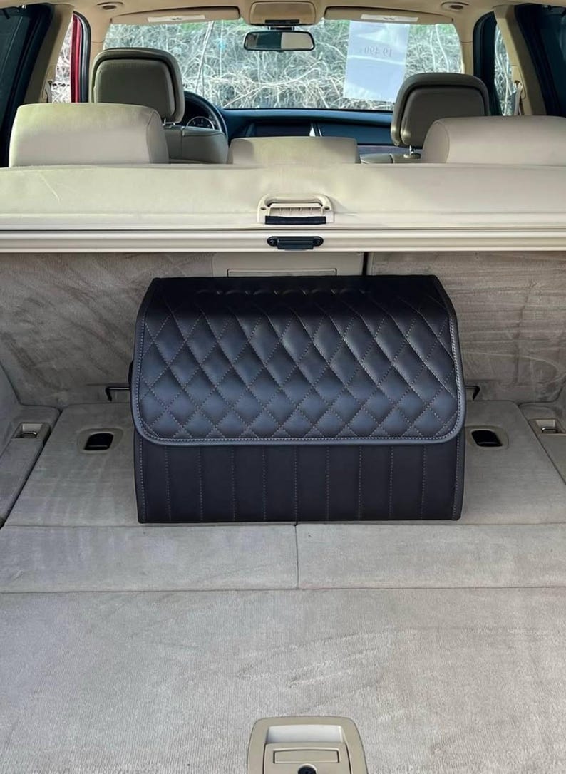 May include: Black quilted car trunk organizer. The organizer is rectangular with a diamond pattern and a top flap. It is designed to fit in the trunk of a vehicle. The interior of the car is visible in the background.