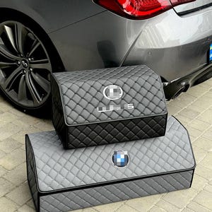 Może przedstawiać: Dwa pikowane organizery do przechowywania w samochodzie, jeden czarny i jeden szary, ułożone przed szarym samochodem. G&oacute;rny organizer ma logo Lexusa, a dolny logo BMW. Oba mają pikowane szwy w romby.