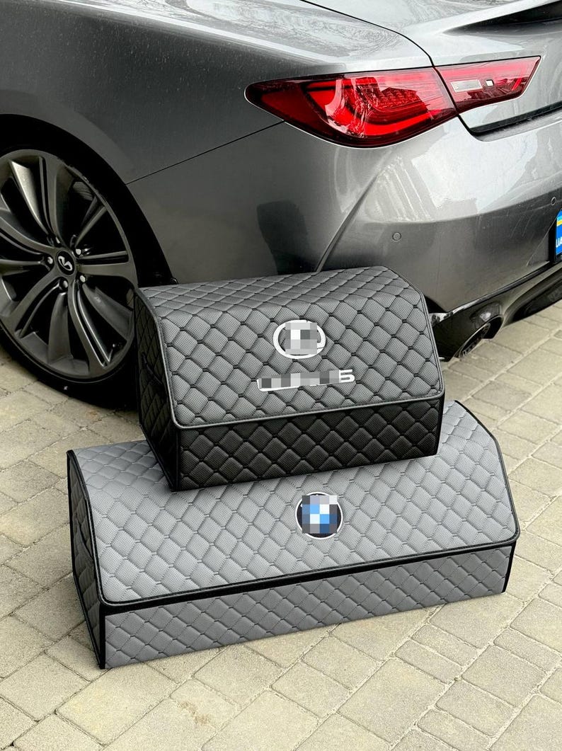 Może przedstawiać: Dwa pikowane organizery do przechowywania w samochodzie, jeden czarny i jeden szary, ułożone przed szarym samochodem. G&oacute;rny organizer ma logo Lexusa, a dolny logo BMW. Oba mają pikowane szwy w romby.