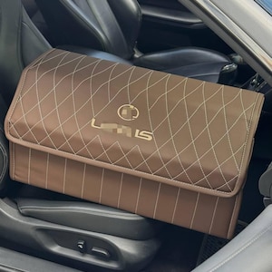 May include: Brown car storage organizer with white diamond stitching and a gold "Lexus LS" logo. The organizer is rectangular and sits on a car seat. The interior of the car is black.