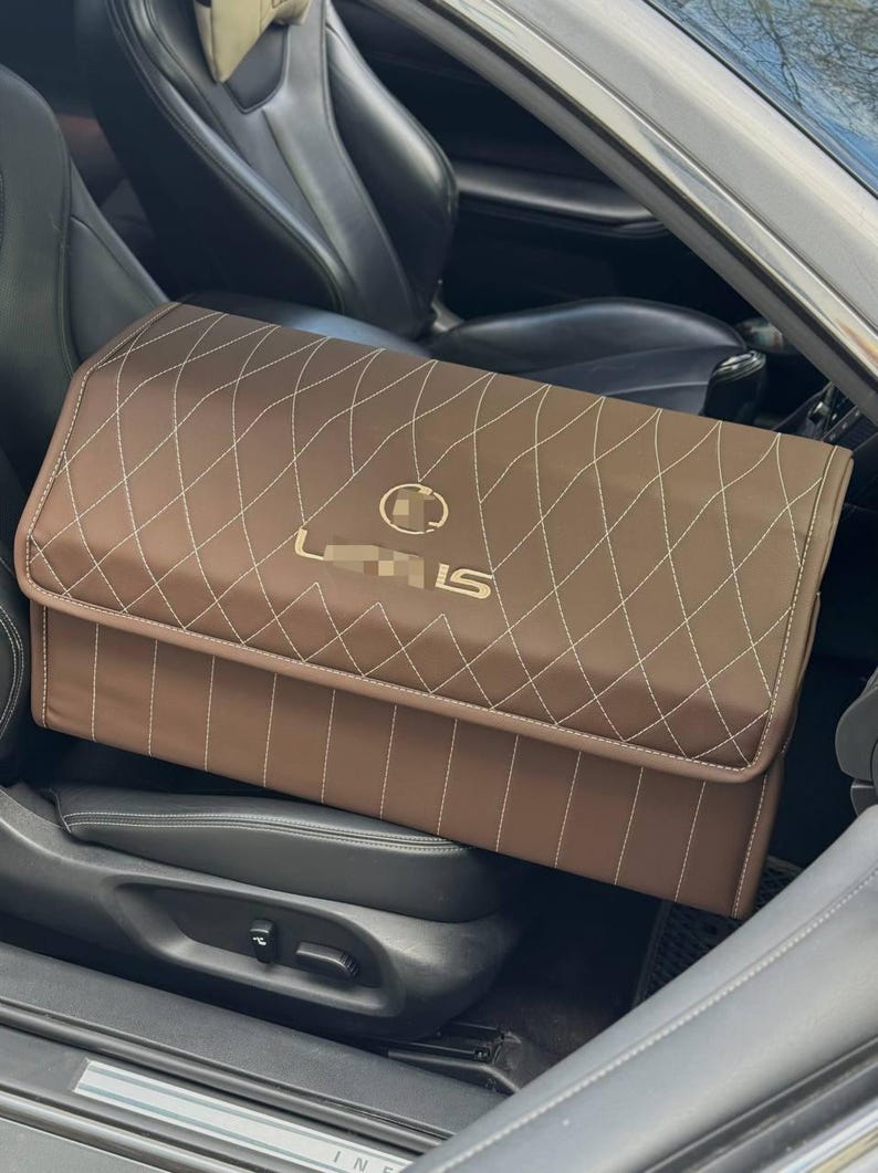 May include: Brown car storage organizer with white diamond stitching and a gold "Lexus LS" logo. The organizer is rectangular and sits on a car seat. The interior of the car is black.