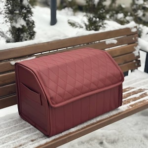 May include: A burgundy-colored, quilted storage box with a curved top and a small side pocket. The box is made of a smooth material and is designed for organizing items. The box is sitting on a wooden bench in a snowy outdoor setting.
