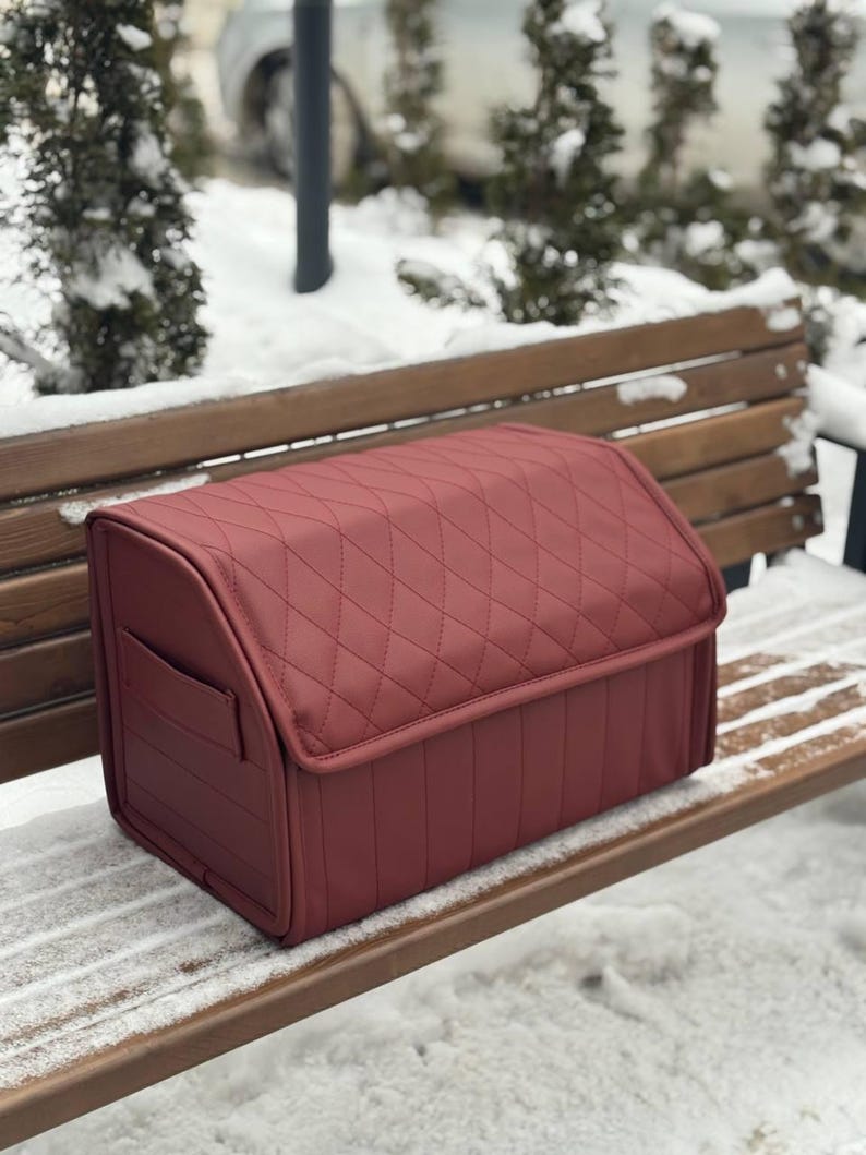 May include: A burgundy-colored, quilted storage box with a curved top and a small side pocket. The box is made of a smooth material and is designed for organizing items. The box is sitting on a wooden bench in a snowy outdoor setting.