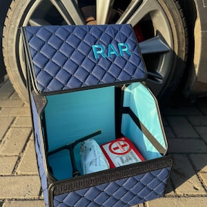 May include: A navy blue quilted car organizer with the letters "RAR" on the lid. The interior is light blue and contains a first aid kit and other items. The organizer is open, revealing its contents.