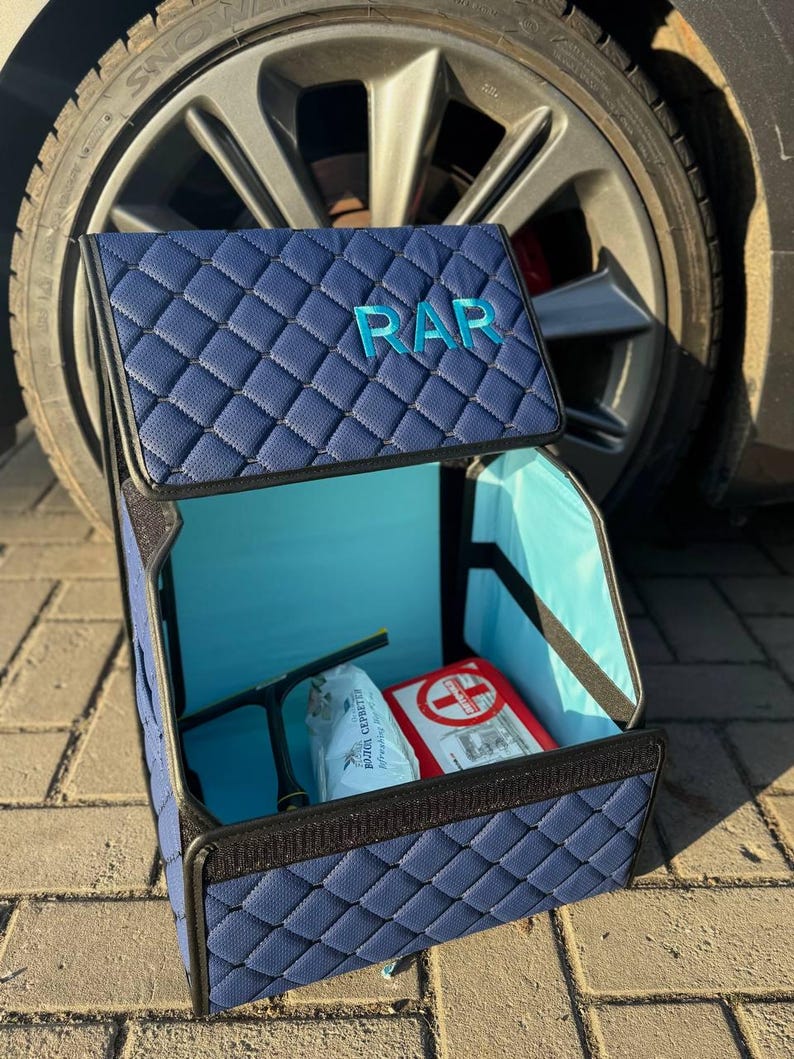 May include: A navy blue quilted car organizer with the letters "RAR" on the lid. The interior is light blue and contains a first aid kit and other items. The organizer is open, revealing its contents.