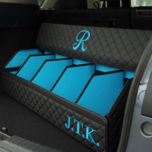 May include: A black quilted car trunk organizer with blue interior compartments. The organizer has the initials "J.T.K." and a stylized "R" embroidered in blue. The organizer is designed to fit in the trunk of a car.
