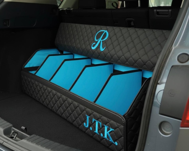 May include: A black quilted car trunk organizer with blue interior compartments. The organizer has the initials "J.T.K." and a stylized "R" embroidered in blue. The organizer is designed to fit in the trunk of a car.