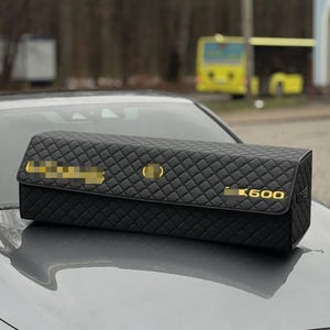 May include: Black quilted car organizer with gold accents and the text "K600". The organizer is rectangular and sits on the hood of a gray car. The background is blurred, showing a bus and trees.
