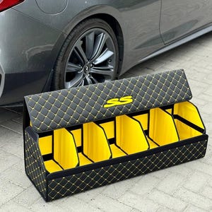 May include: Black and yellow quilted car trunk organizer with multiple compartments. The organizer has a diamond pattern with yellow stitching and a yellow interior. The logo "SS" is visible on the lid. A gray car is in the background.