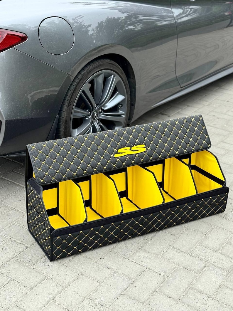 May include: Black and yellow quilted car trunk organizer with multiple compartments. The organizer has a diamond pattern with yellow stitching and a yellow interior. The logo "SS" is visible on the lid. A gray car is in the background.