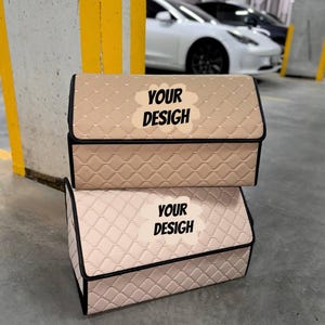 May include: Two beige car storage organizers with a quilted pattern and black trim are stacked on top of each other. Each organizer has a lid and the text "YOUR DESIGN" on the front. The background shows a parking garage with cars.