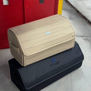May include: Two car storage organizers. The top one is tan with a geometric design and a small blue logo. The bottom one is black with a blue logo. Both are rectangular with rounded edges and handles.