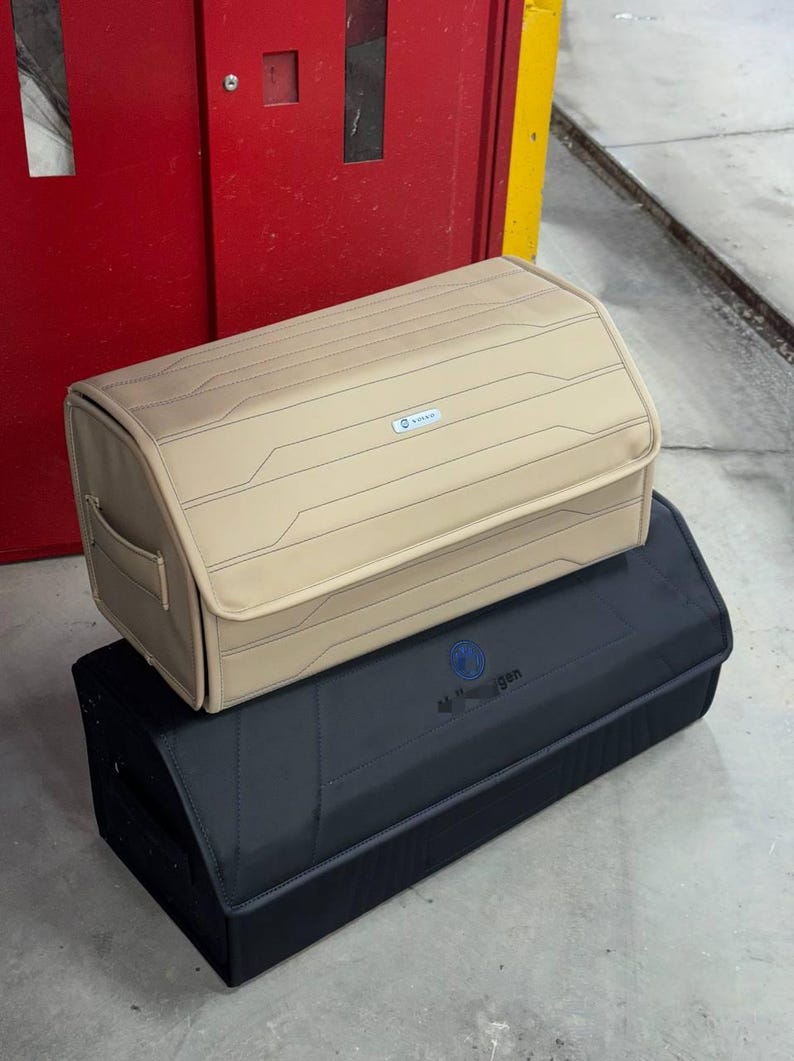 May include: Two car storage organizers. The top one is tan with a geometric design and a small blue logo. The bottom one is black with a blue logo. Both are rectangular with rounded edges and handles.
