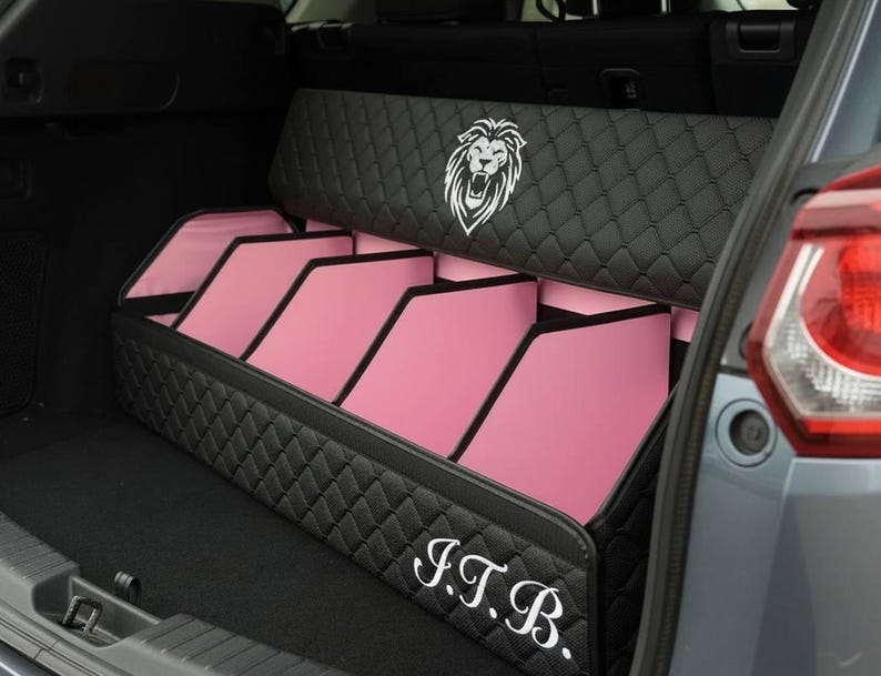 May include: Black quilted car trunk organizer with pink interior compartments. Features a white lion emblem and the initials "J.T.B." in script. The organizer is designed to fit in the trunk of a vehicle.