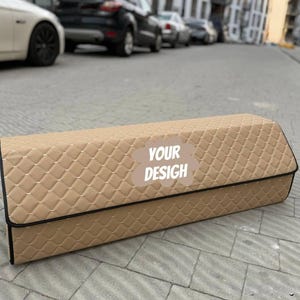 May include: A tan, rectangular car trunk organizer with a diamond-quilted pattern. The organizer has a black trim and the text "YOUR DESIGN" on the front. The organizer is sitting on a gray, paved surface.