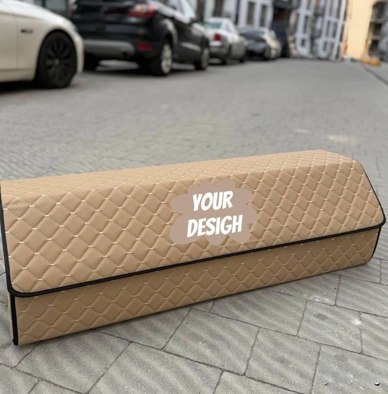 May include: A tan, rectangular car trunk organizer with a diamond-quilted pattern. The organizer has a black trim and the text "YOUR DESIGN" on the front. The organizer is sitting on a gray, paved surface.