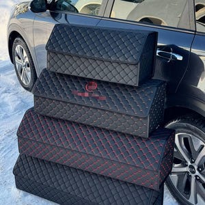 May include: Black car trunk organizers with a quilted pattern. The organizers are stacked, with red stitching on one. The car is parked in the snow.
