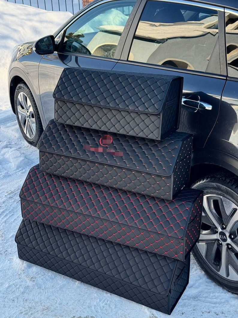 May include: Black car trunk organizers with a quilted pattern. The organizers are stacked, with red stitching on one. The car is parked in the snow.
