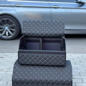May include: Black car trunk organizer with a quilted diamond pattern and white stitching. The organizer is shown in two stacked sections, with the top section open to reveal interior compartments. A silver car is in the background.