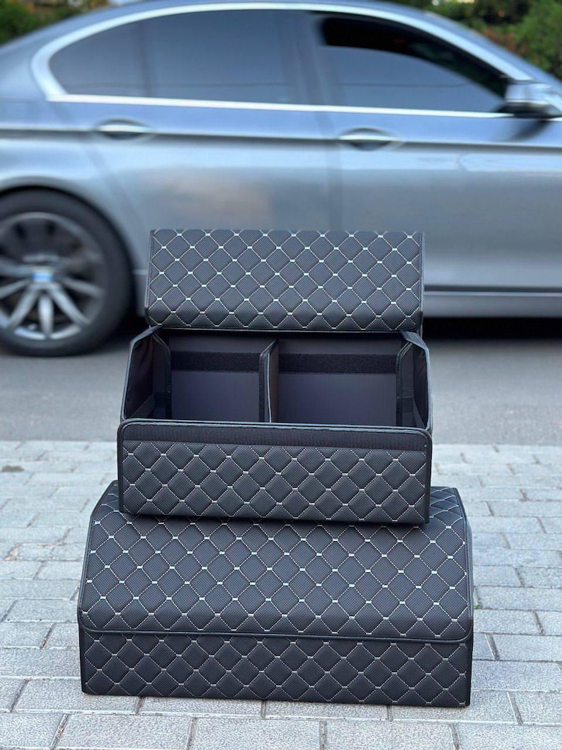 May include: Black car trunk organizer with a quilted diamond pattern and white stitching. The organizer is shown in two stacked sections, with the top section open to reveal interior compartments. A silver car is in the background.