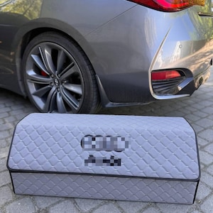 May include: A gray car trunk organizer with a quilted pattern. The organizer is rectangular with a black trim and is positioned in front of a gray car. The car has a red taillight.